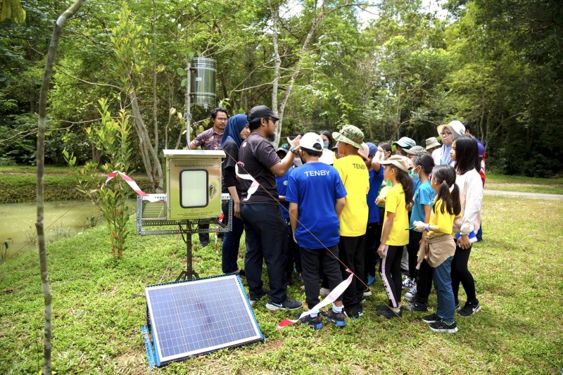 A large group of schoolchildren gather round water and rain level monitoring equipment on a riverbank as they listen to Hub researchers
